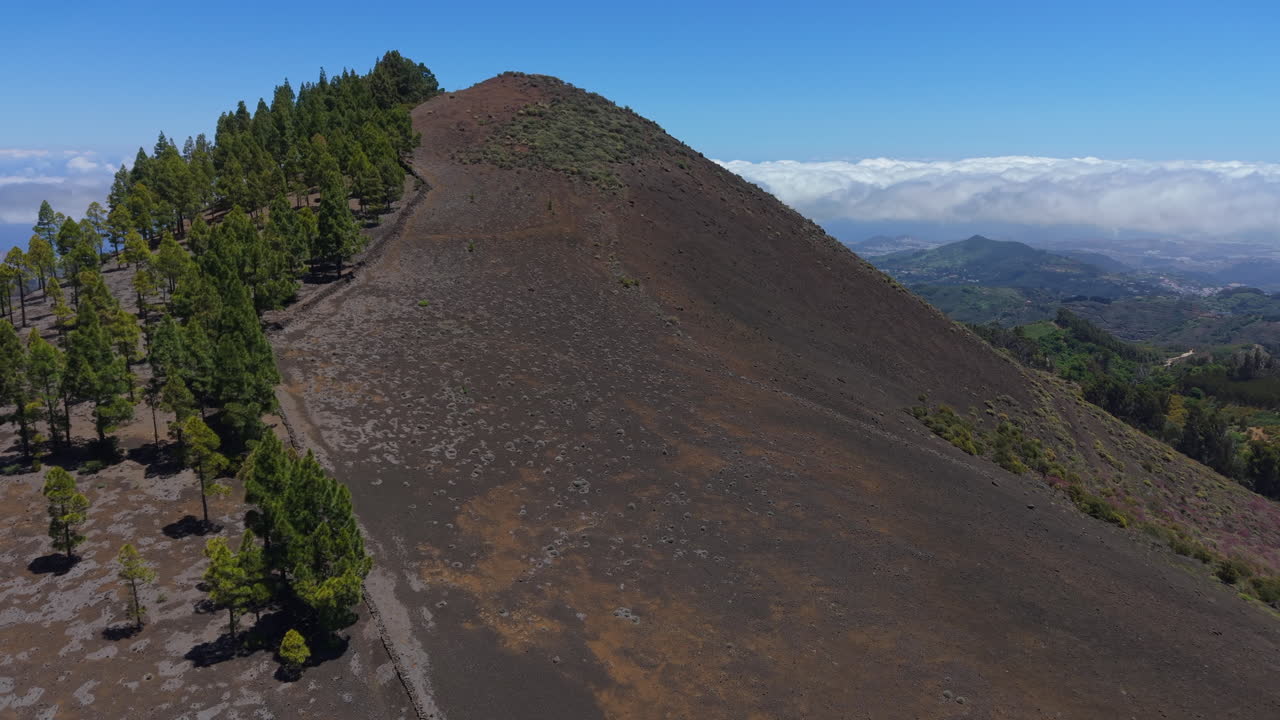 Dramatic black volcanic mountain (Montañon Negro) rising majestically through cloud layers, revealing rugged Gran Canaria landscape under bright sunlight with lush vegetation contrasting rocky terrain