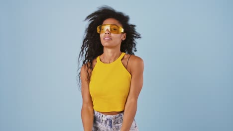 afro-american model in yellow sunglasses is smiling, dancing and shaking her long curly hair while posing on blue background. slow motion