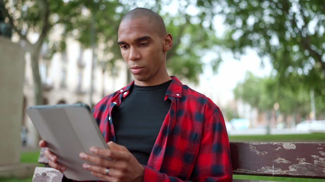 Serious bald African man typing on digital tablet on bench
