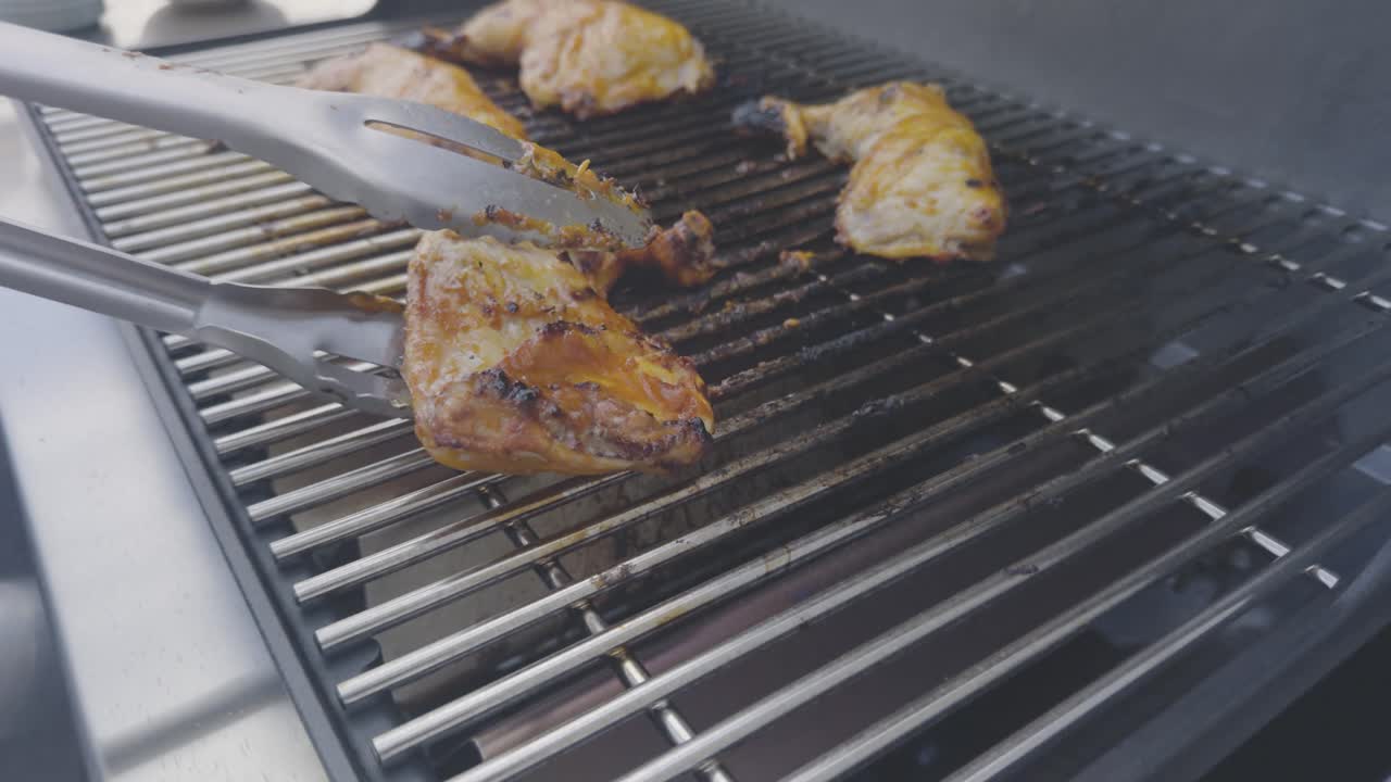 Grilled chicken legs cooking on a stainless steel BBQ grill. Stainless steel tongs turn the chicken as the camera pulls back on an angle across the grill