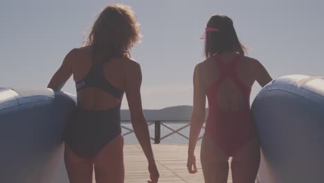 medium back view shot of two good looking girls holding paddle boards in hands getting ready for surfing