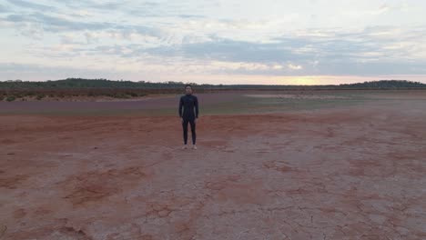 drone clip moving towards male model with dark hair wearing wetsuit, stood still in red desert landscape