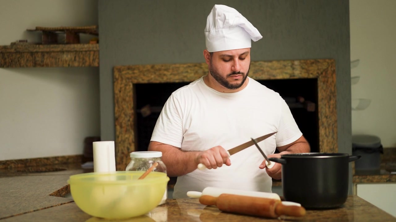 Uno chef esperto affila con cura un coltello su un acciaino, preparandosi per la preparazione del pasto con vari ingredienti disposti su un bancone.
