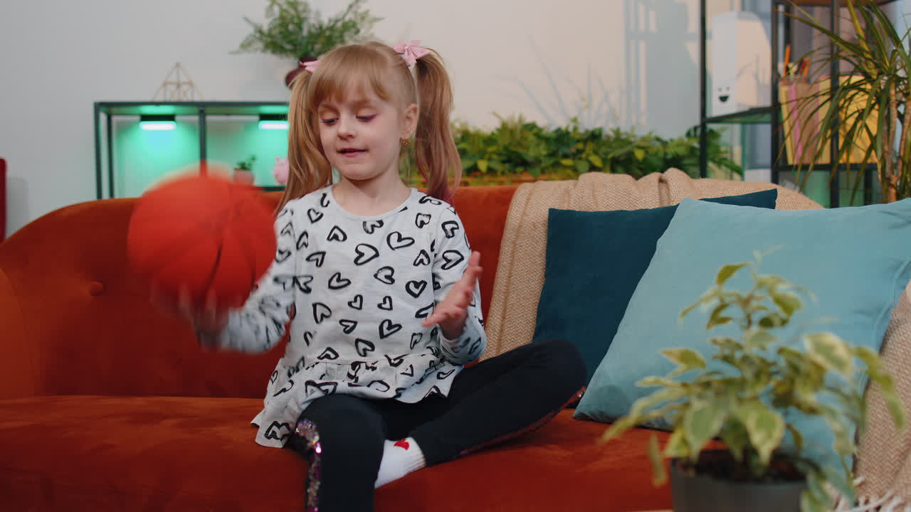Young little children girl basketball fan holding ball looking at camera, training dribbling at home