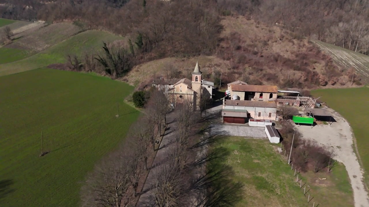 La chiesa di Sant'Ilario Vescovo si affaccia su Chiavenna Rocchetta, Lugagnano Val d'Arda, Piacenza, Emilia-Romagna, Italia, tra campi coltivati e colline boscose, sorvolo in avanti del drone in discesa