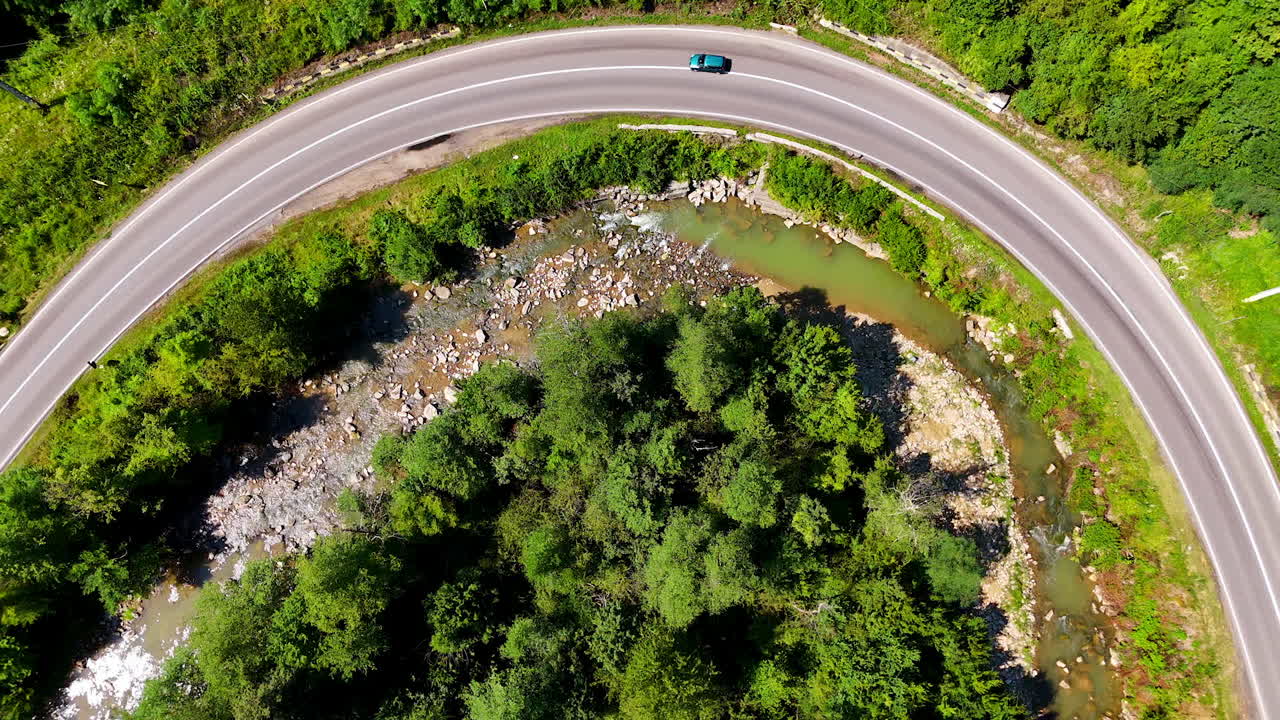 Winding road circles green area near flowing stream. A car travels along a winding road amidst lush green foliage and a clear stream, creating a serene natural atmosphere