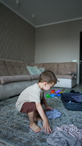 joyful caucasian toddler throws his clothes to the ceiling. a suitcase and scattered toys on the floor at backdrop. vertical video.
