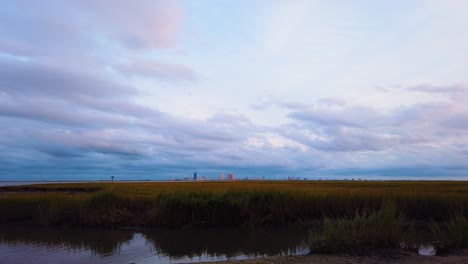 4K-TIMELAPSE-Atlantic-City-skyline-in-distance-over-waterway-with-mostly-cloudy-sky-day-to-night-with-clouds-turning-pink,-purple-and-blue