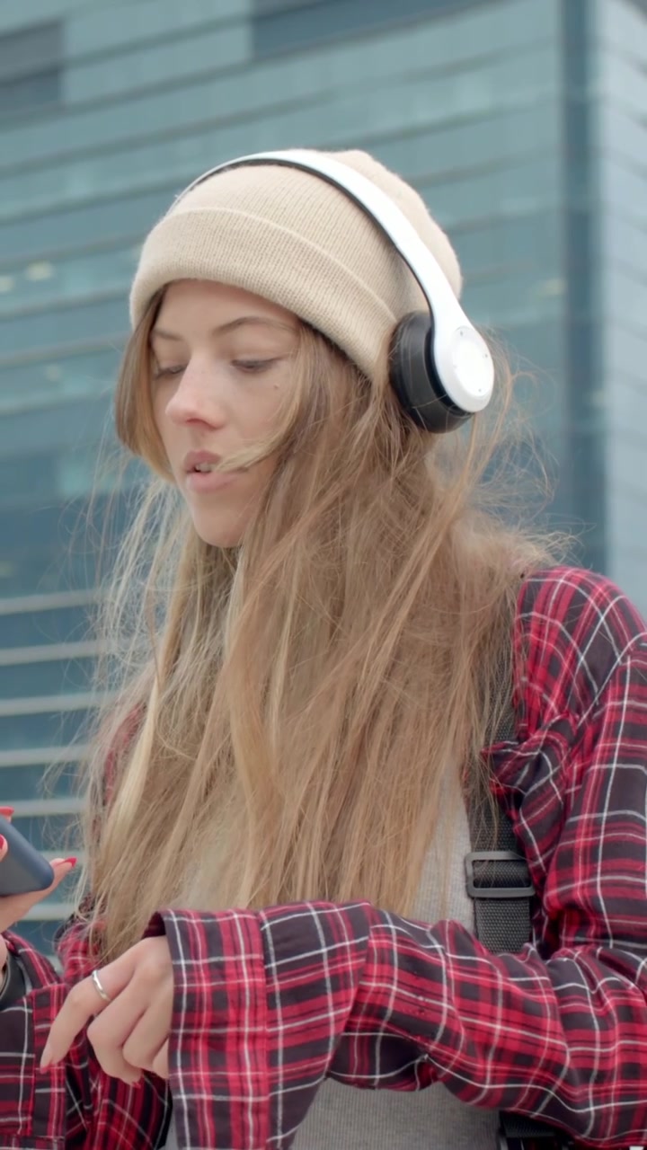 Video verticale: Vento che fa sì che una donna con il berretto tenga le cuffie per stabilizzarle e controlli il telefono vicino a una parete di vetro.