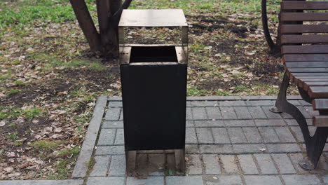 metal trash can positioned beside wooden bench on paved path in peaceful urban park, surrounded by fallen leaves and green grass, emphasizing cleanliness and outdoor public space atmosphere