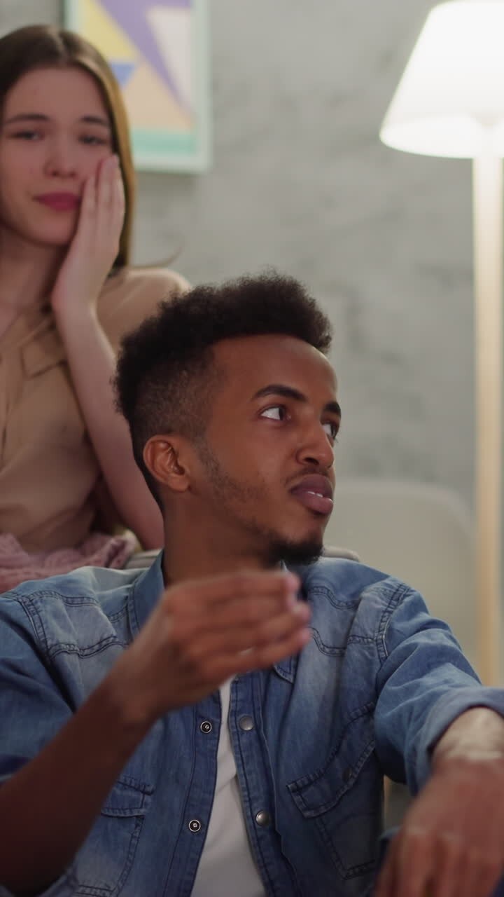 Black man talks to guy companion sitting near blonde girlfriend covered with plaid at home closeup slow motion. Young people at cinema evening in apartment