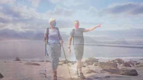 pareja de ancianos caucásicos con máscaras faciales caminando por la costa, sobre el mar en movimiento