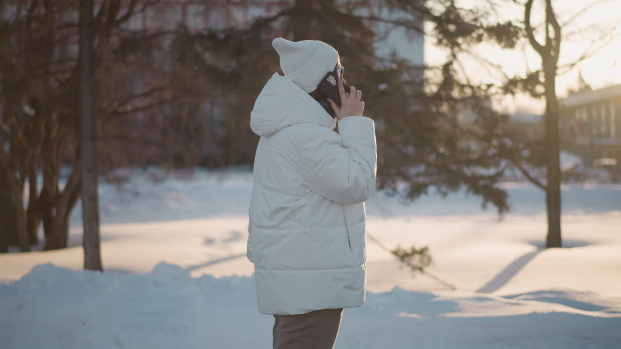 Free spirit holds phone to ear singing and dancing in rhythm under sunset light wearing beanie and puffer coat near statue and snowy trees backdrop conveying joyful vibe with subtle silhouette effect
