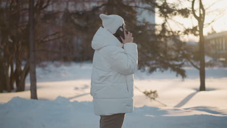 free spirit holds phone to ear singing and dancing in rhythm under sunset light wearing beanie and puffer coat near statue and snowy trees backdrop conveying joyful vibe with subtle silhouette effect