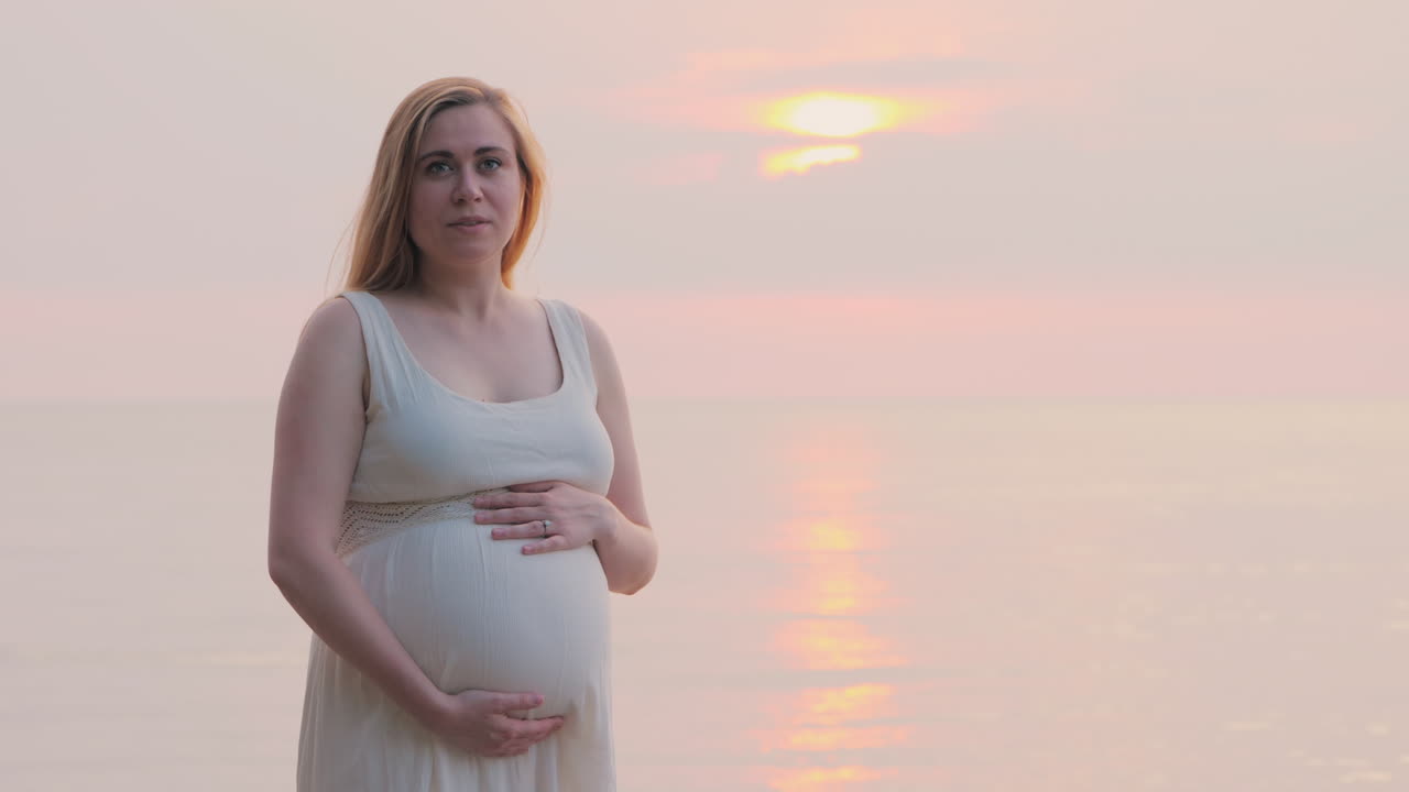 임신한 여자는 그녀의 배를 쓰다듬어 그녀의 침대 뒤에 바다 근처에 서있다