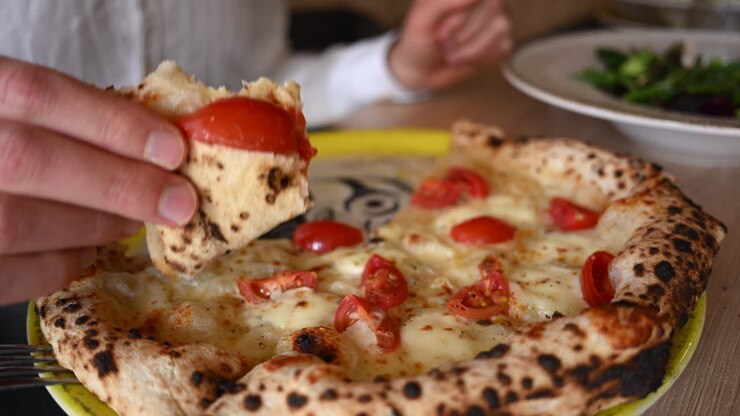 Mano che solleva una fetta di pizza al formaggio appena sfornata e condita con pomodorini.