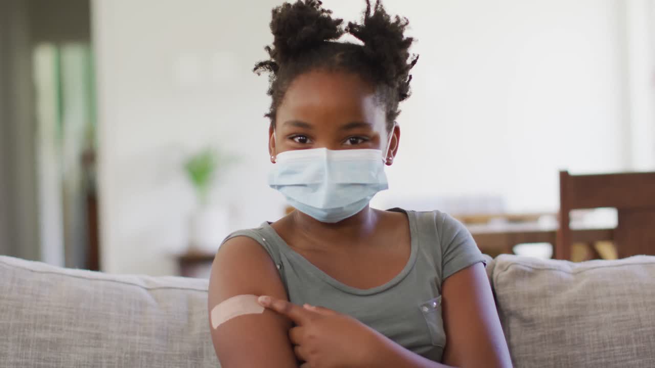 Afroamerikansk flicka som bär ansiktsmask pekar på plåster på armen