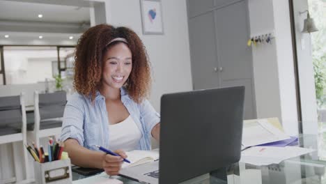 Portrait-of-happy-biracial-woman-using-laptop-and-writing-notes-at-home,-in-slow-motion