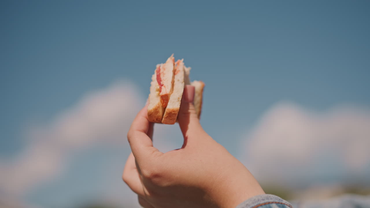 Mano che tiene un panino contro il cielo blu, primo piano illuminato dal sole mostra un toast ripieno di marmellata tenuto da una mano vestita di jeans, nuvole soffici e toni caldi evocano un'atmosfera rilassata da picnic, le briciole evidenziano la consistenza e l'aspetto casalingo