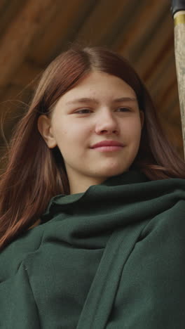 smiling girl in dark green cloak stands in gazebo. young female warrior holds spear and waits for fighting training on blurred background close low angle shot