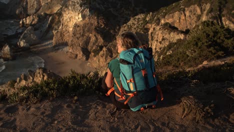 rear view of woman with backpack sitting at cliff