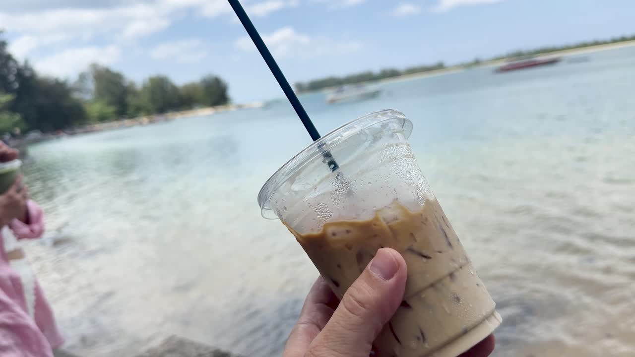Hand holds iced coffee by clear ocean, sunny beach, gentle camera movement, relaxed mood