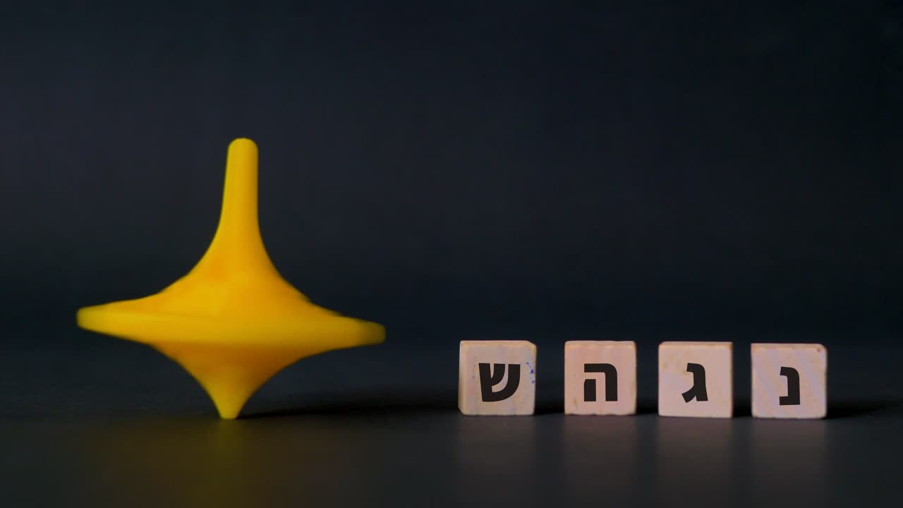 una peonza de plástico de hanukkah gira junto a las cuatro letras hebreas nun, gimel, heh y shin que se muestran sobre un fondo negro