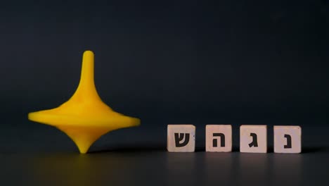 A-plastic-Hanukkah-spinning-top-spins-beside-the-four-hebrew-letters-nun,-gimel,-heh-and-shin-shown-against-a-black-background