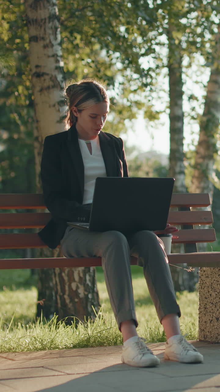 Freelancer takes break drinking water while working outdoors with laptop on lap, seated on wooden bench in park with sunlight filtering through trees, surrounded by lush greenery