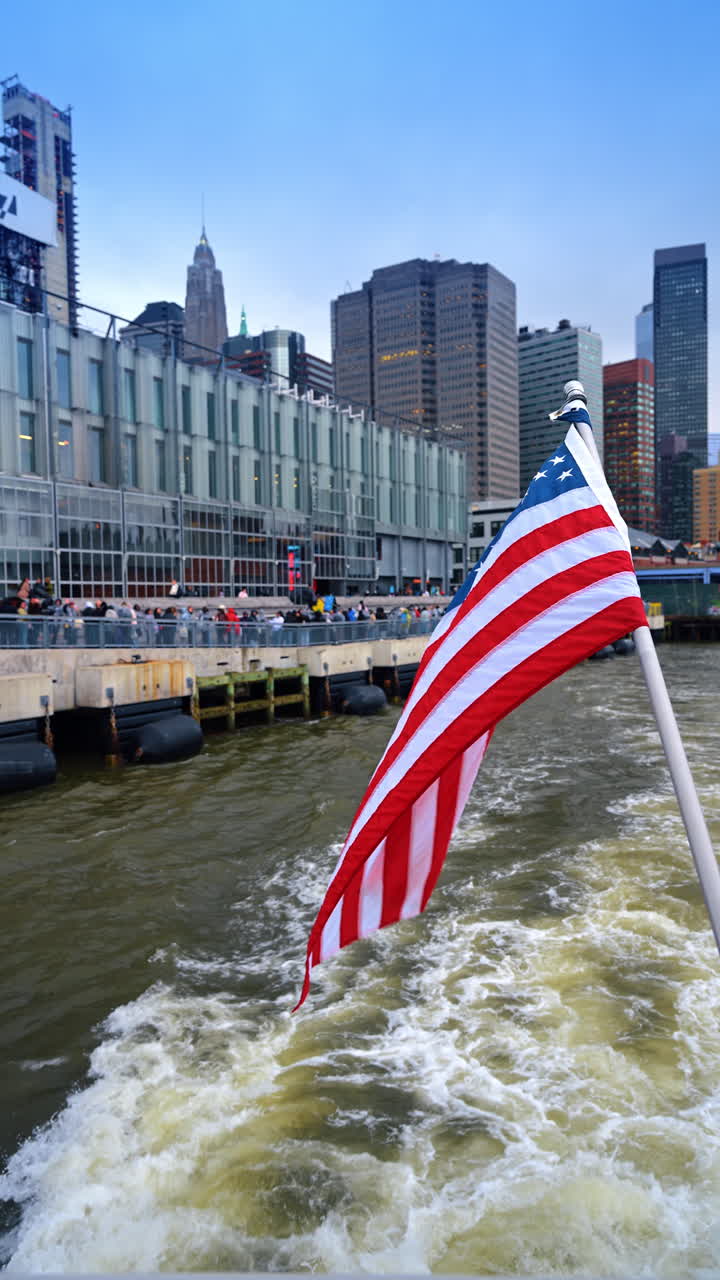 Traghetto che lascia il Molo 17 con bandiera americana e skyline. Vista dal retro della barca che mostra la scia dell'acqua, la bandiera USA e i grattacieli di Lower Manhattan a New York.