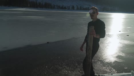 Man-breaking-ice-along-shore-of-frozen-mountain-lake-with-sunlight-reflected-on-icy-surface