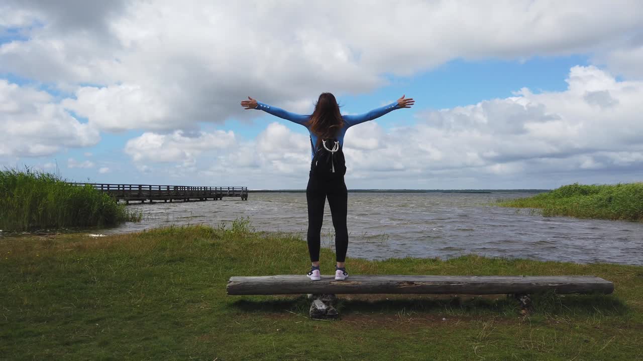 Young woman standing on a wooden bench with open arms by a lake in Poland, enjoying freedom and nature