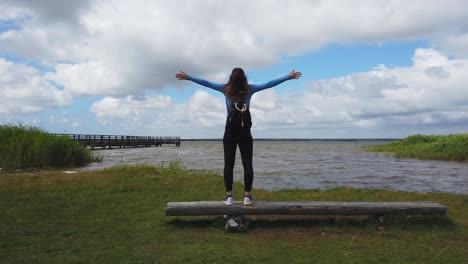 Young woman standing on a wooden bench with open arms by a lake in Poland, enjoying freedom and nature