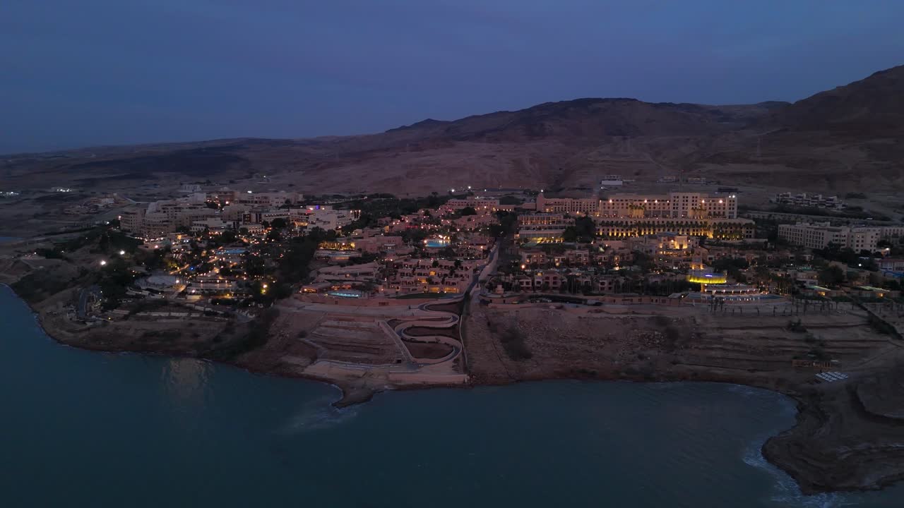 Ürdün'deki Ölü Deniz kıyıları boyunca uzanan lüks bir tatil köyü kompleksini, sakin mavi saatte sıcak ışıklarla aydınlatılmış olarak gösteren havadan drone çekimi