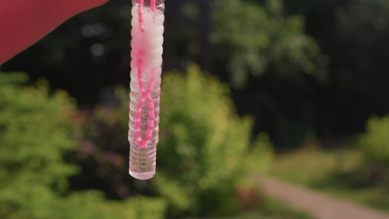 CloseUp White Woman Bead Droplet Hanging From Textured Tube Outdoors, Tiny Pink Beads And Clear Gel Form Slow Motion Pendant At Tip, Sunlight Catches Facets Against Soft Green Bokeh, Hypnotic ASMR