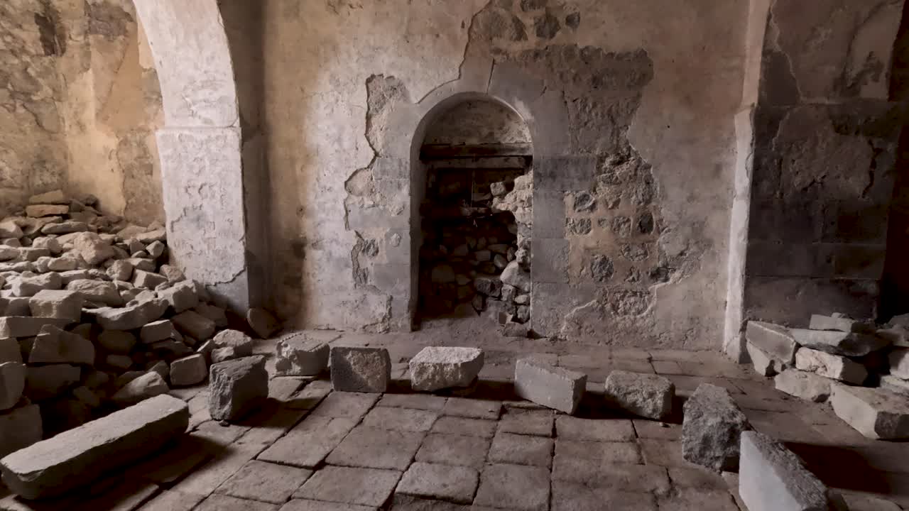 Dentro de uma igreja antiga, pedras estão espalhadas pelo chão. Uma pintura mural pode ser vista. A luz entra pelas janelas, revelando a estrutura antiga. Este lugar guarda história.