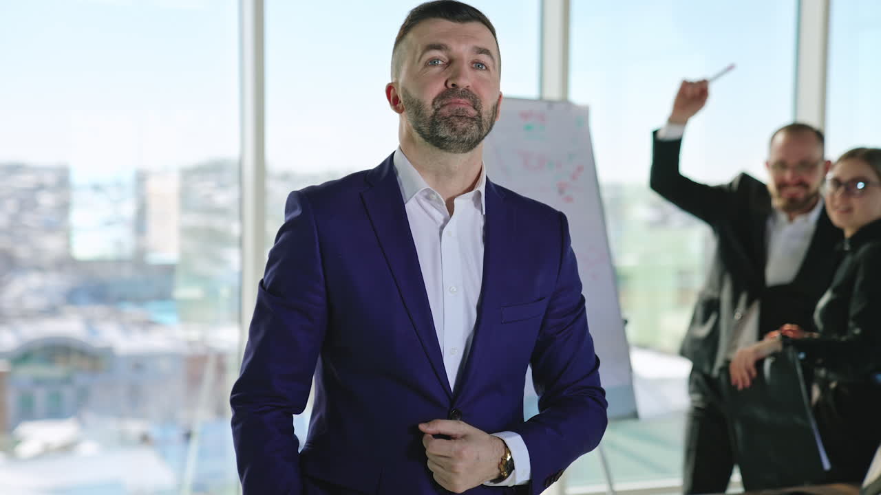 Businessman wearing blue suit standing in a big office. Man of business talking about something in front of the camera. Two people from office staff at the background.