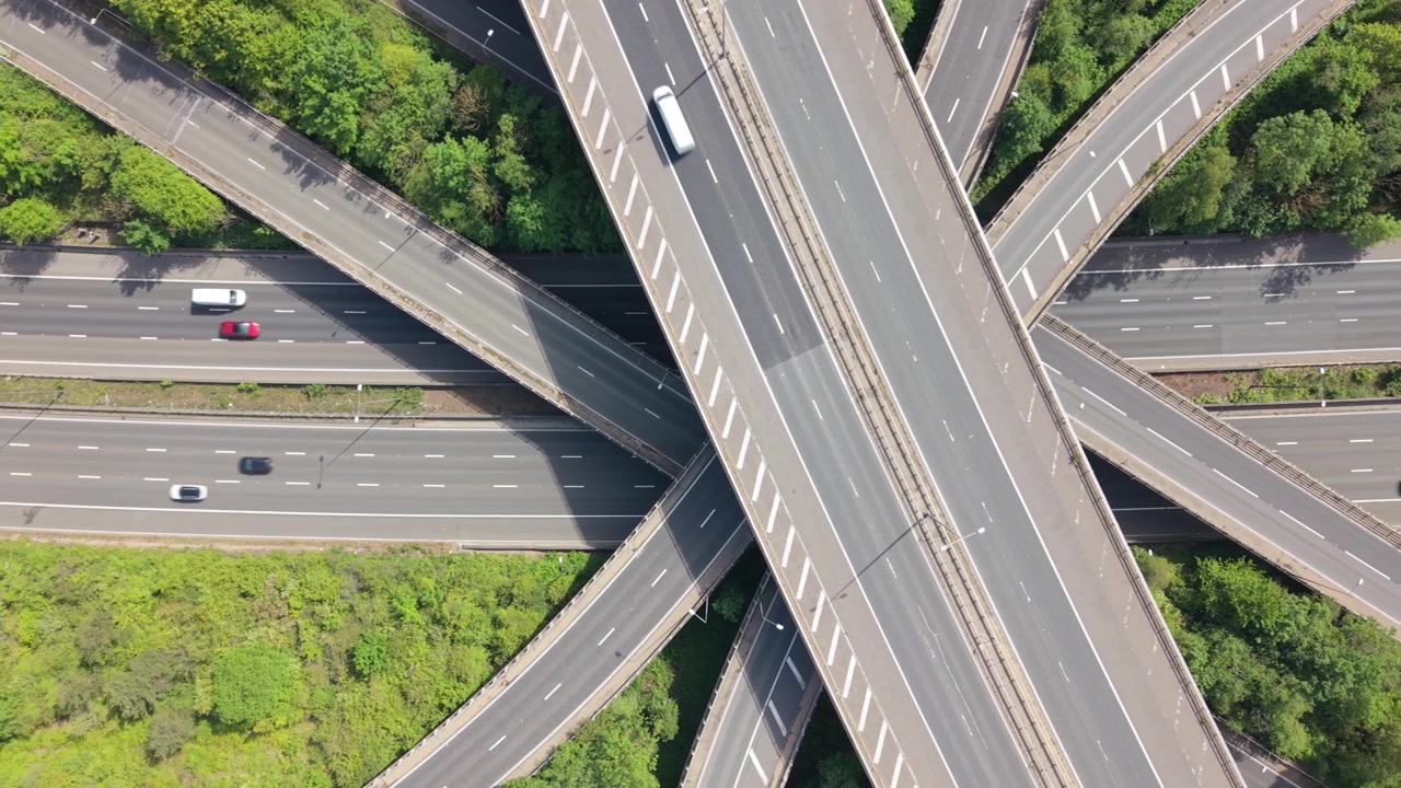 Overhead view captures vehicles traversing a massive, multi-level highway interchange. Automobiles and trucks drive on intersecting roads amidst lush green vegetation during a bright day.
