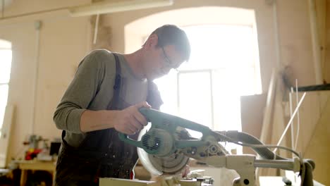 medium shot side view of mature furniture maker using circular saw to make cuts in wooden plank in workshop lit with sunlight