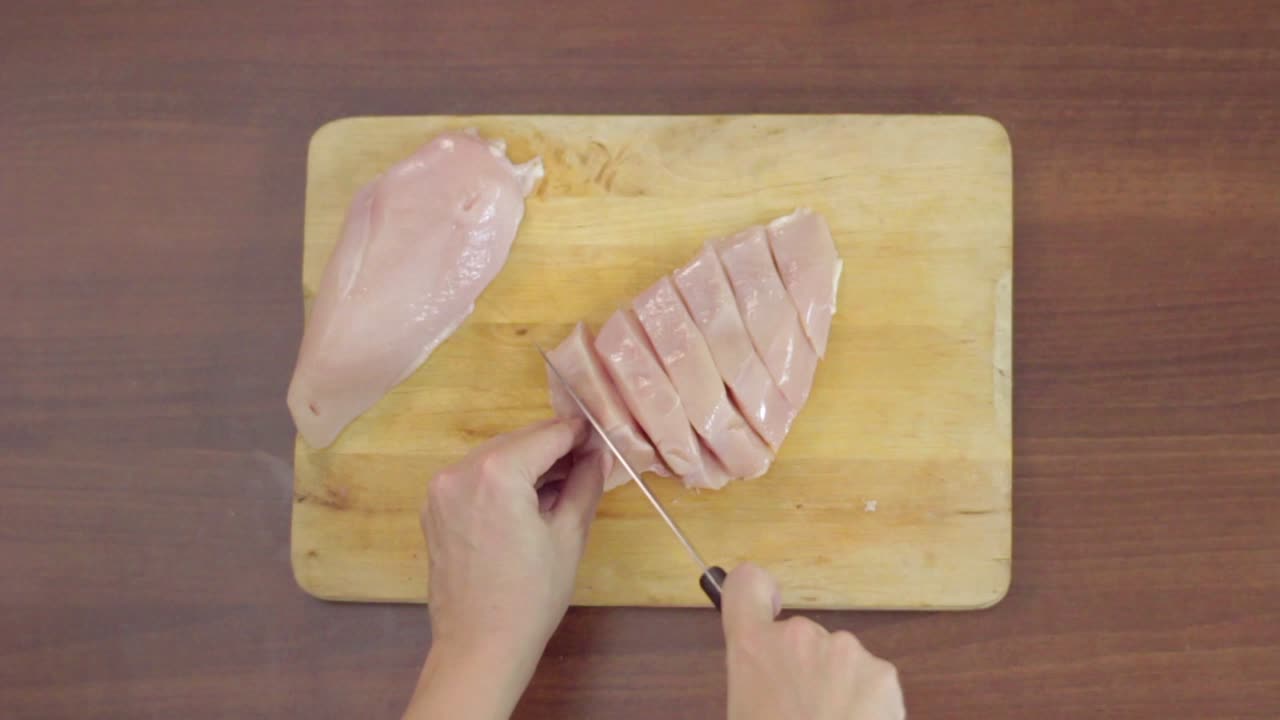 Mani che tagliano pollo crudo a cubetti su un tagliere di legno