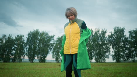 mujer en chaqueta amarilla bajo un impermeable verde se quita su impermeable en un campo de hierba brumosa bajo un cielo nublado con árboles en la distancia