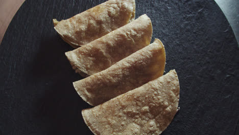 Wunderschöne-Zeitlupenaufnahme-Einer-Gruppe-Von-4-Tacos-Oder-Mexikanischen-Tortillas,-Während-Die-Kamera-Leicht-Um-Den-Braunen-Teller-Rotiert-Und-Die-Tortillas-Mit-Einem-Schwachen-Weißen-Licht-Beleuchtet-Werden