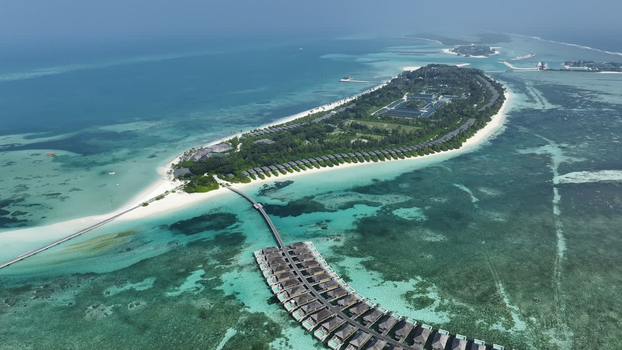 Ampia veduta aerea con drone che mostra una fila curva di lussuose ville sull'acqua collegate da una passerella che conduce a un'isola resort lussureggiante e verde con una barriera corallina visibile e isole distanti nell'oceano profondo