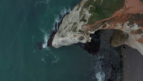 Slow-Epic-Birds-Eye-Top-Down-View-Etretat-Cliffs-on-Overcast-Day-with-dark-blue-ocean,-waves-crashing-on-rocks