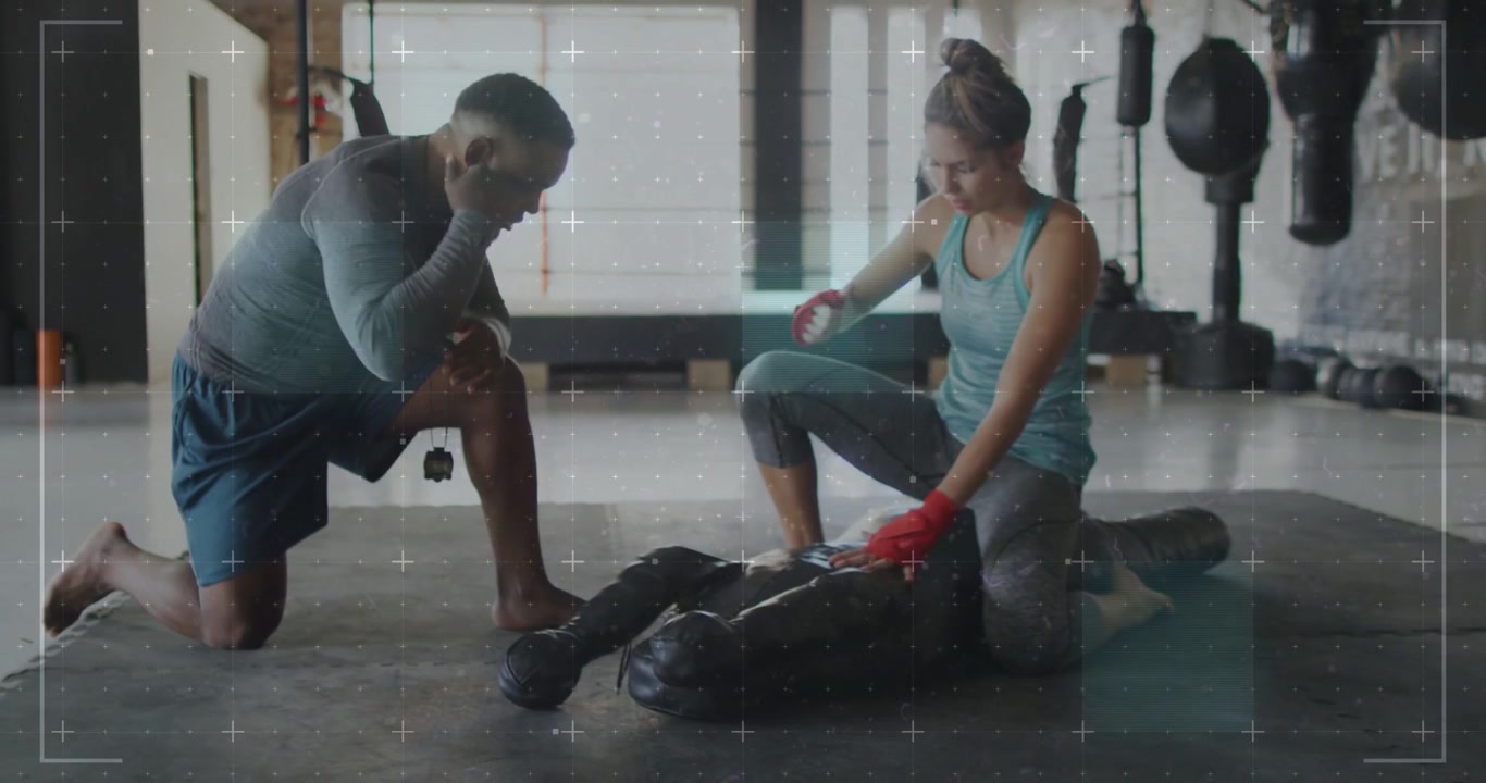 Male trainer cueing female trainee kneeling on bag practicing punches AR showing red wraps for gym