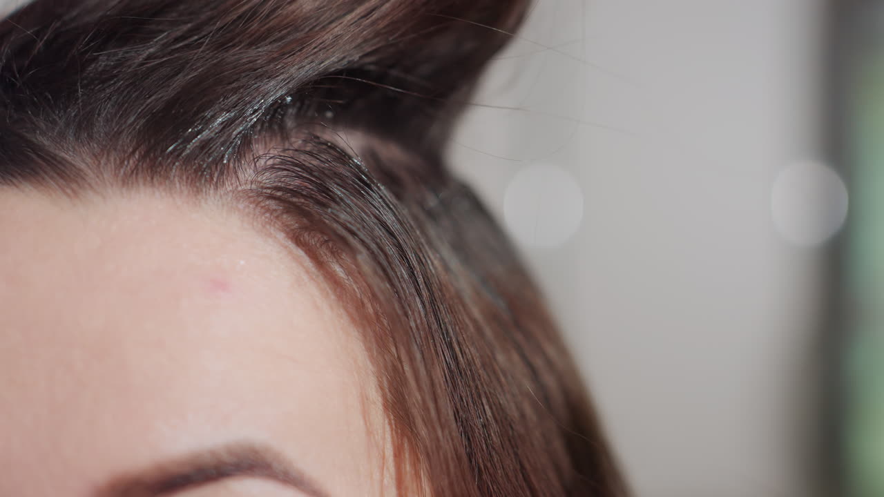 브러시로 자라난 머리카락에 염색약을 바르고, 스타일리스트가 머리카락 섹션을 들어 올리고, 정확하게 가르마를 나누고, 두피 디테일까지 보여주는 클로즈업 헤어라인 뿌리 염색 수정, 차분한 미용실 거울 배경과 전문적인 기술에 초점을 맞췄어요.