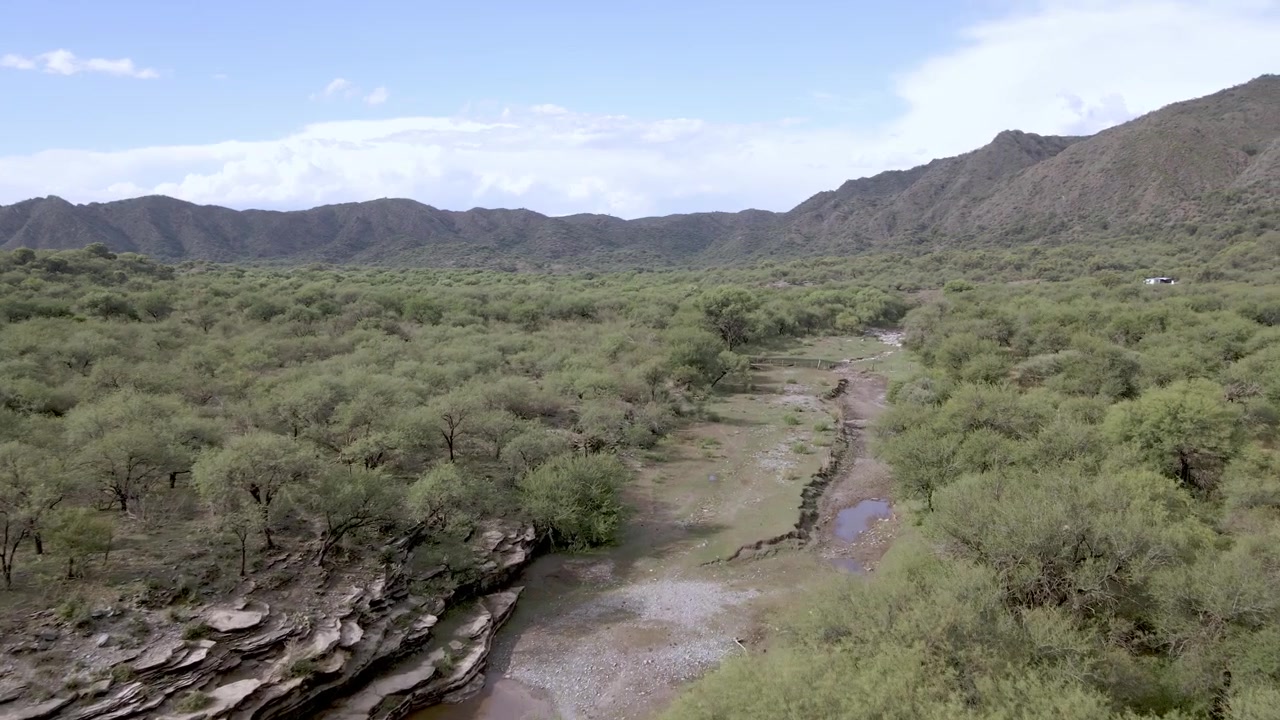 Margens rochosas em camadas, um leito de rio raso e sinuoso e uma densa floresta de arbustos verdes situam-se abaixo de montanhas baixas no Parque Provincial Bajo de Véliz, Argentina.