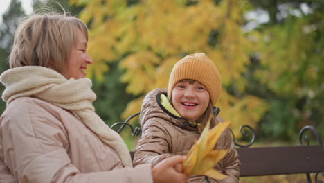 child holding bright yellow autumn leaf excitedly jumps onto mother sitting on park bench, sharing affectionate hug and warm peck amid golden foliage and cool breeze