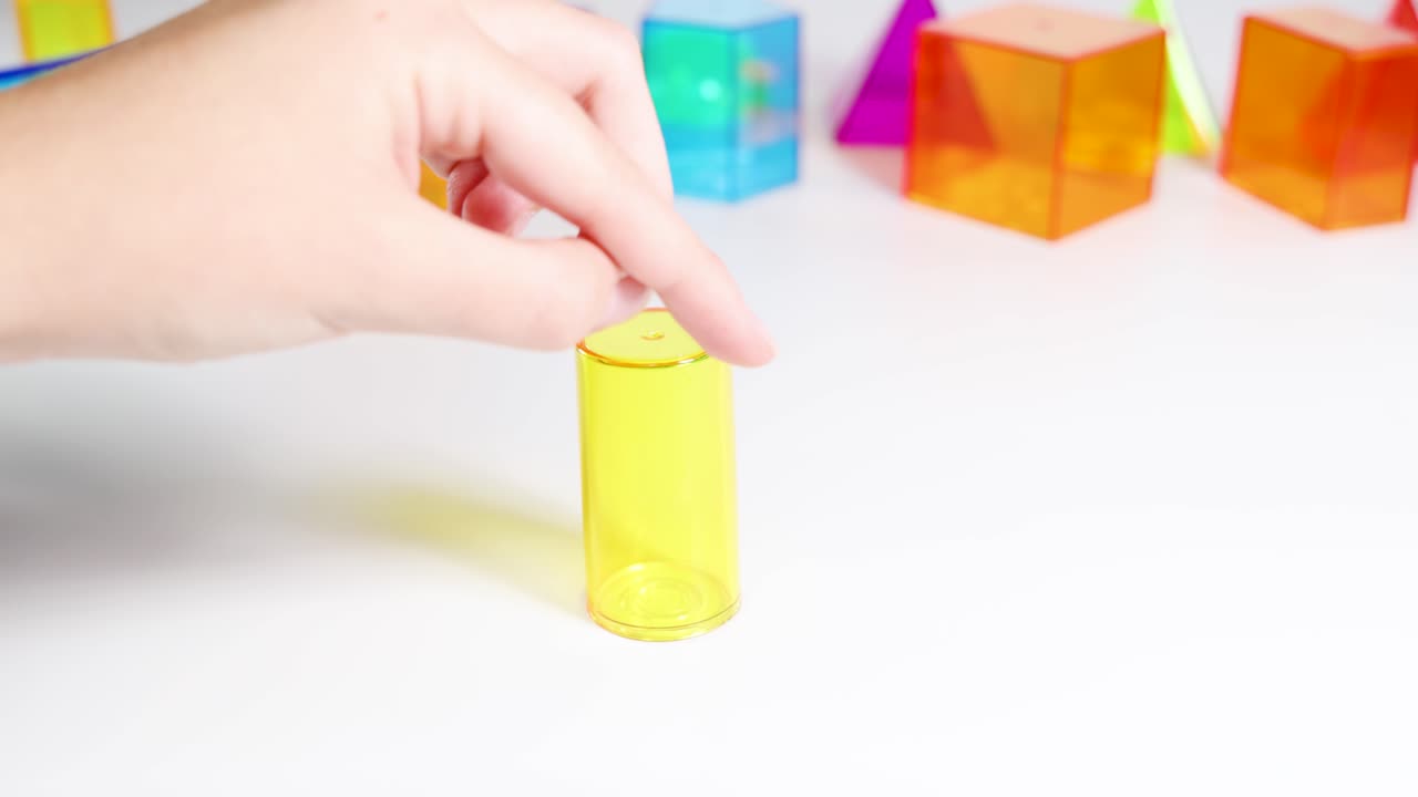 Hands manipulate vibrant 3D geometric shapes on a white surface, showcasing educational interaction with prisms and cylinders