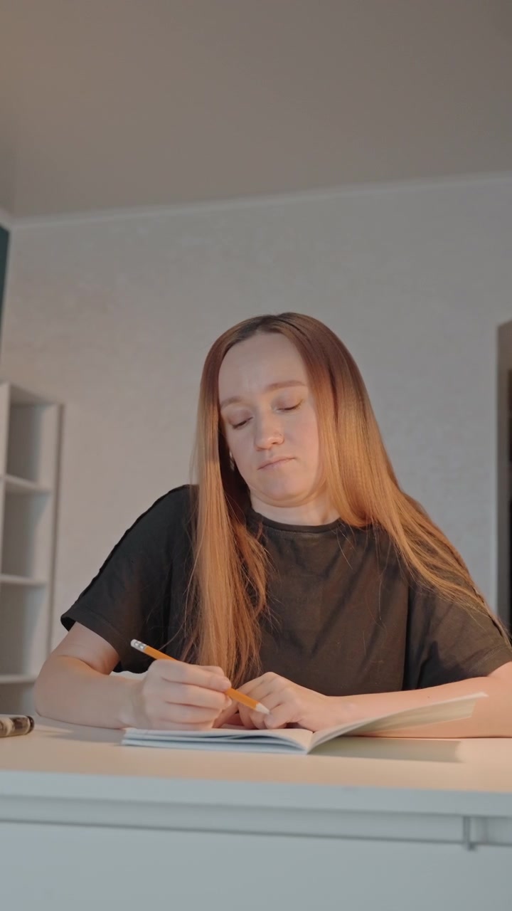 Giovane donna scrive su un quaderno con una matita arancione. La donna indossa una maglietta nera e ha lunghi capelli castano chiaro, due penne. L'ambiente interno presenta pareti bianche e scaffali pieni